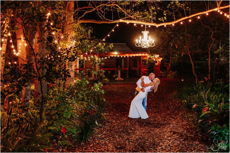 groom dips bride and kisses her under outdoor chandelier and market lights surrounded by trees at rustic barn wedding