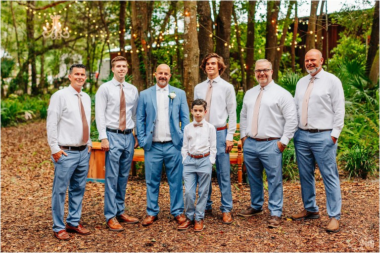 groom and groomsmen standing under market lights before rustic barn wedding in light blue and pale pink