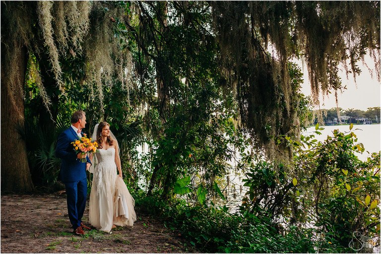bride and groom look out at lake as they stroll through the massive oaks at Kraft Azalea park wedding