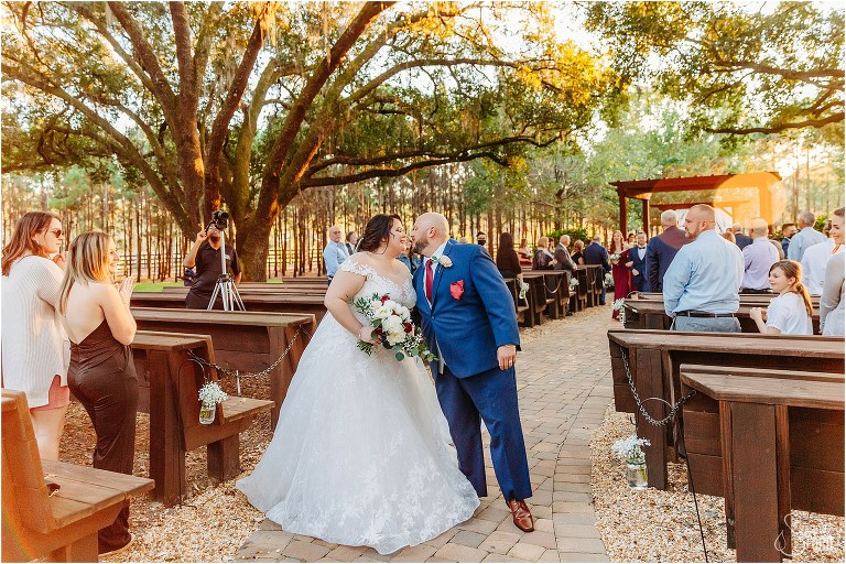 bride and groom kiss at end of aisle as loved ones cheer at rustic Apopka wedding