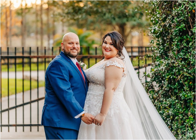 bride and groom hold hands and cheese for the camera in front of wrought-iron gate and ivy arch
