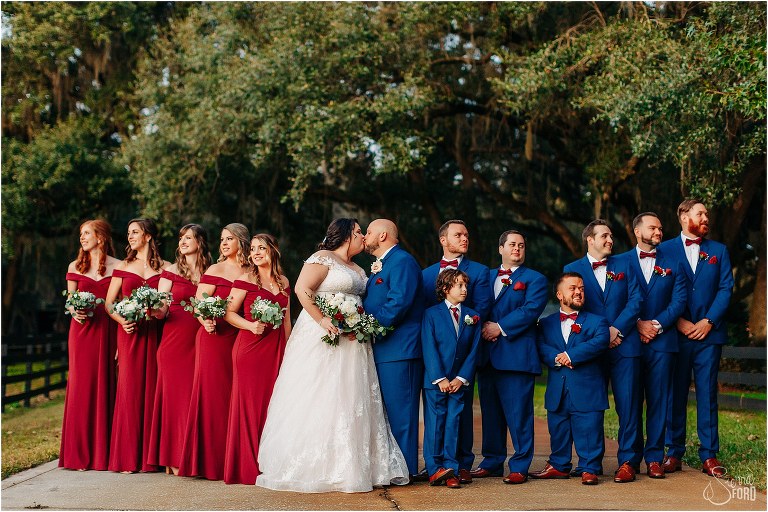 couple kisses as wedding party look off in opposite directions in navy and wine-colored outfits at rustic Apopka wedding
