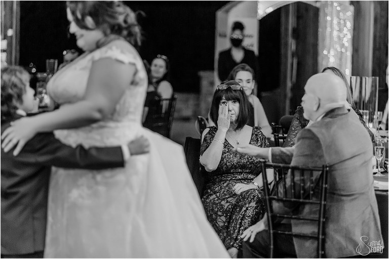 mother of bride wipes tears from her eyes while bride dances with son
