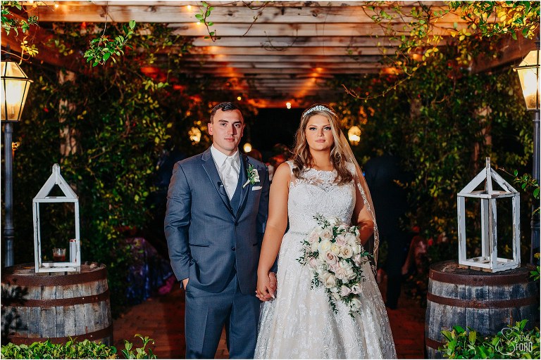 bride & groom stand hand in hand under garden market lights at Hidden Barn wedding