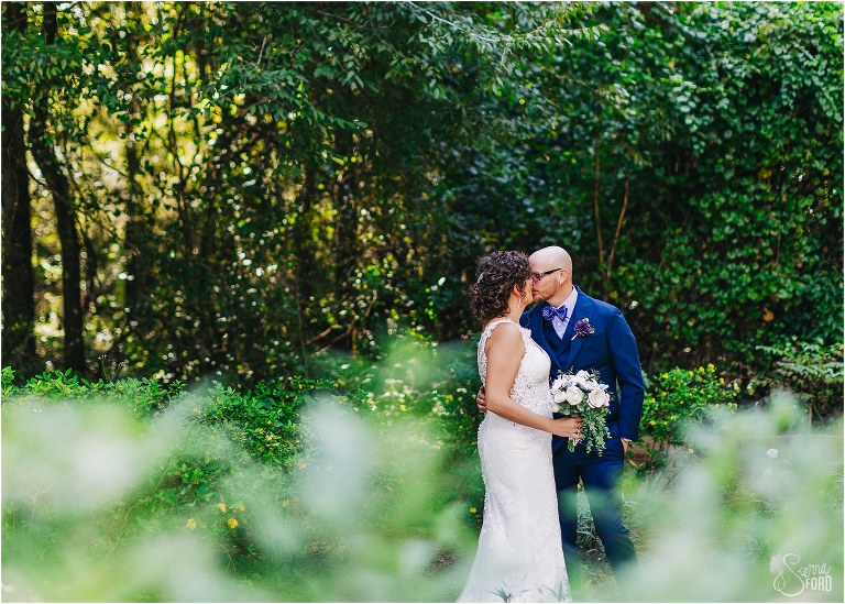 groom sneaks a kiss with bride as camera peeks through bushes at October Oaks wedding