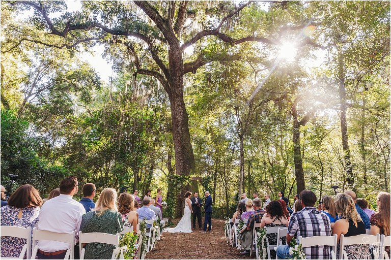 sun streaks through giant oak trees as bride & groom exchange vows at October Oaks wedding