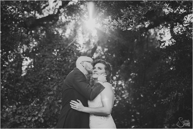black and white of sun streaming through leaves as groom kisses bride's cheek at October Oaks wedding